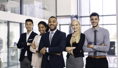 A diverse group of five young professionals confidently pose together in a modern office setting. They are smiling and dressed in business attire, conveying teamwork and success. The spacious and well-lit environment reflects a contemporary and dynamic professional space.